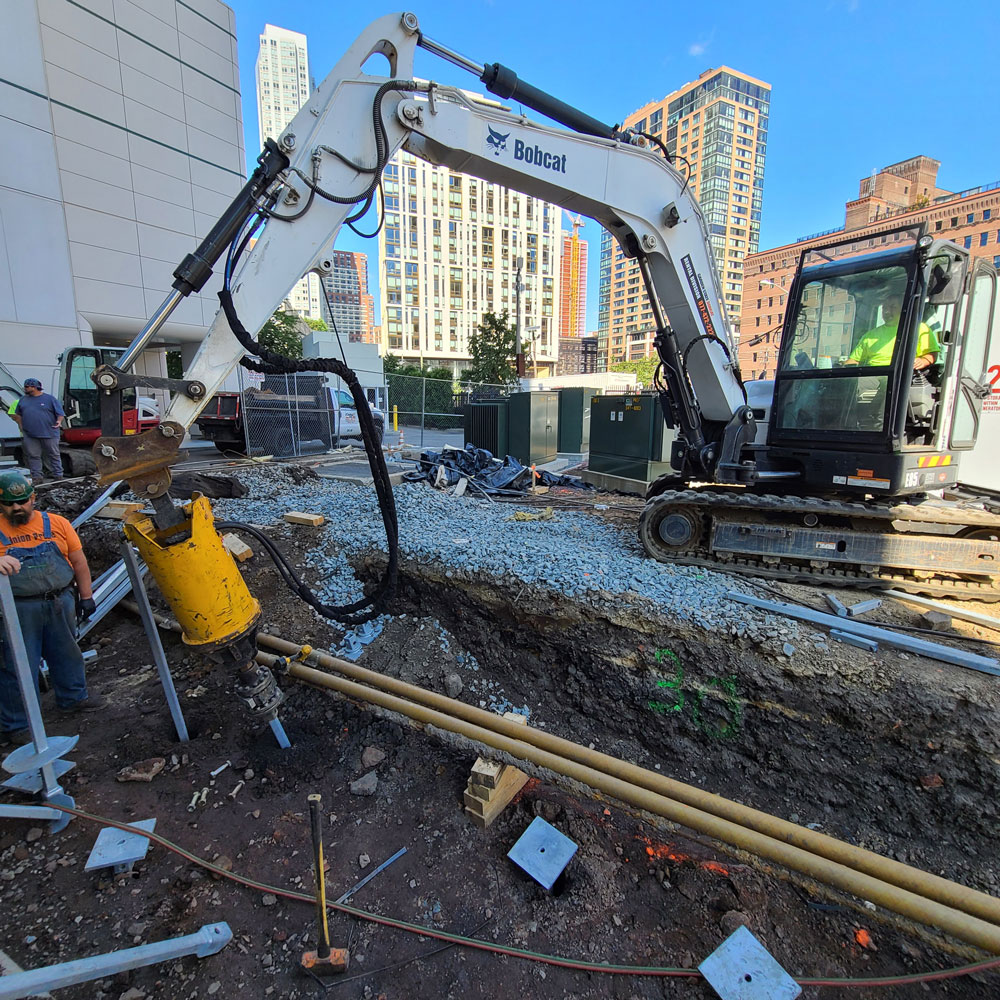 worker standing by while bobcat drives a helical pile into the concrete flood wall foundation